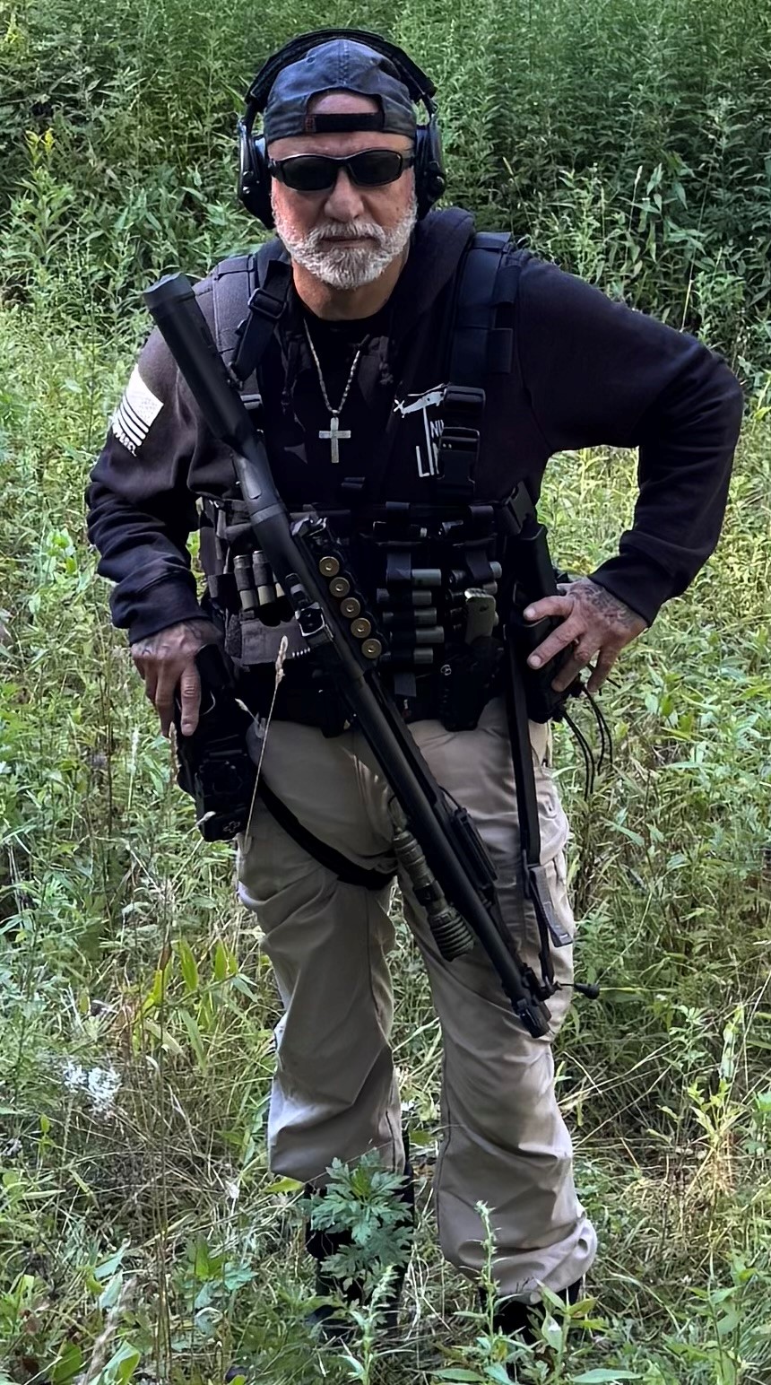 Joe Falcone, former NYPD officer, in tactical gear with a shotgun, standing in a grassy area, emphasizing his commitment to law enforcement and mortgage assistance for police families.